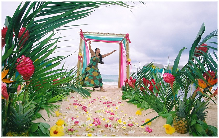 Hula dancer in a green floral dress raising her arms under a pink and turquoise draped arch, with a petal-strewn aisle and tropical flowers
